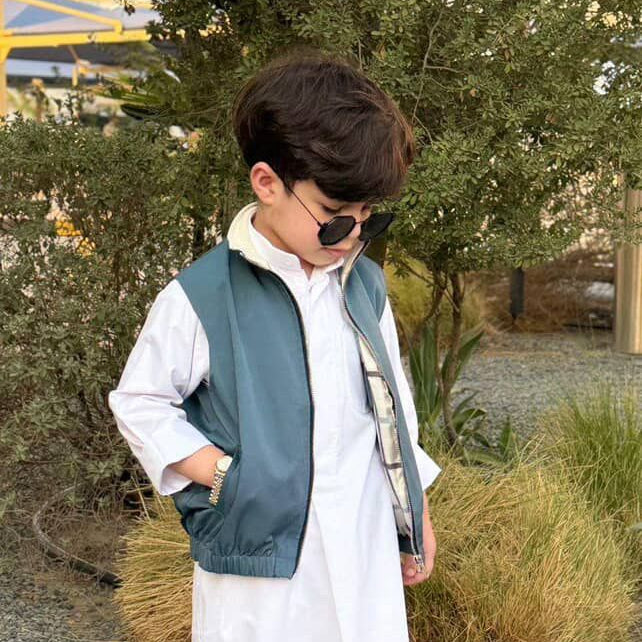 Child wearing a long white coat, blue vest, and sunglasses outdoors.